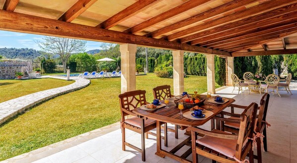 A covered outdoor dining area with a stone pathway leading to a pool, surrounded by lush greenery and a stone wall.