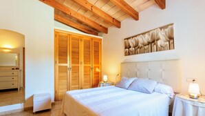 A bedroom with a large bed, wooden ceiling beams, a wardrobe, and a wall-mounted artwork.