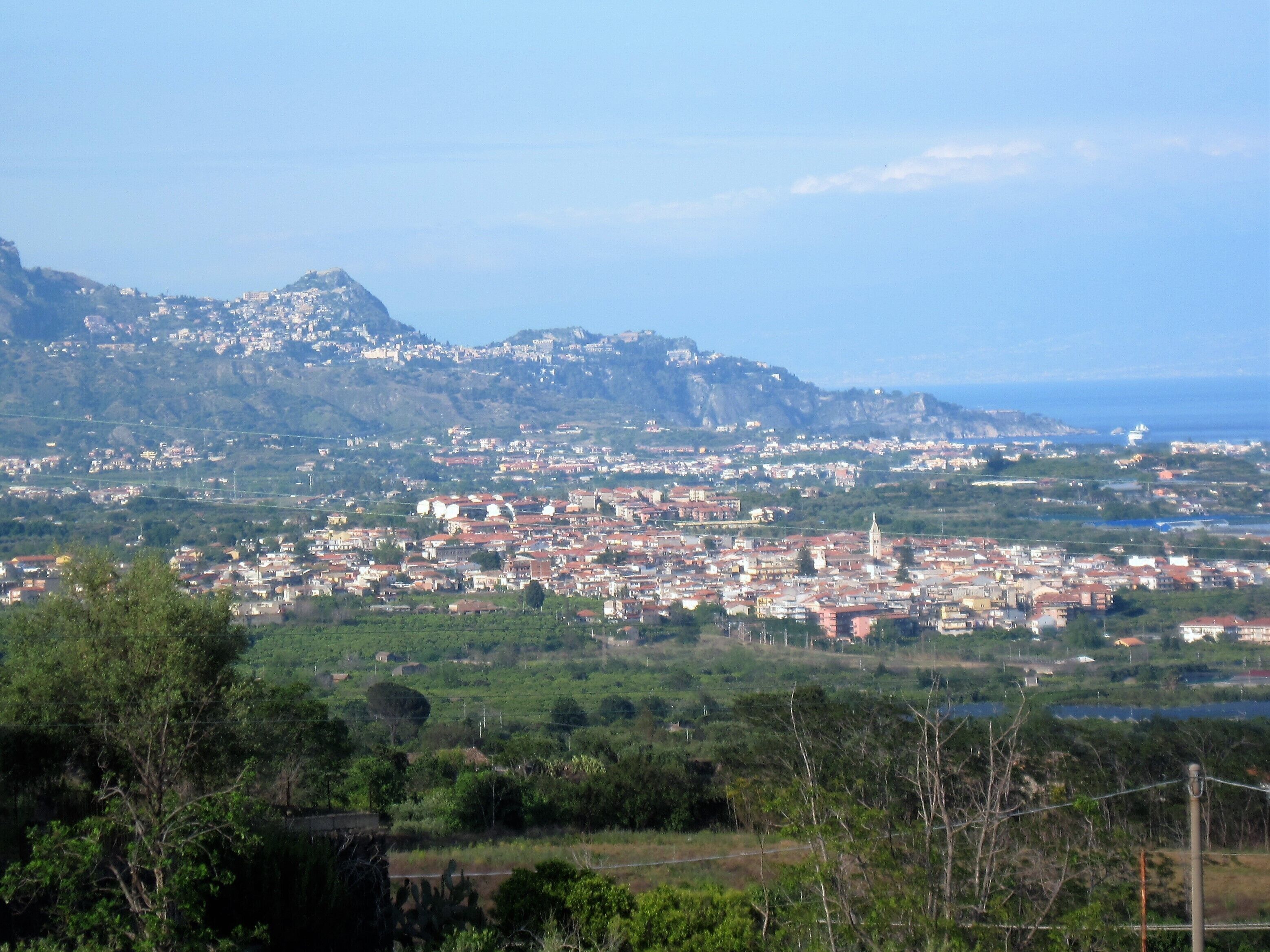 Paesaggio costiero con una montagna imponente sullo sfondo, zona urbana densa con edifici dai tetti rossi e una distesa d'acqua in lontananza.