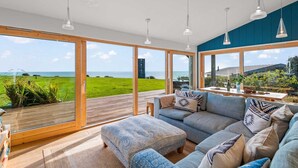 Amplio salón con un gran ventanal corredera que da a un área exterior con césped, una pared de acento azul y una terraza de madera.