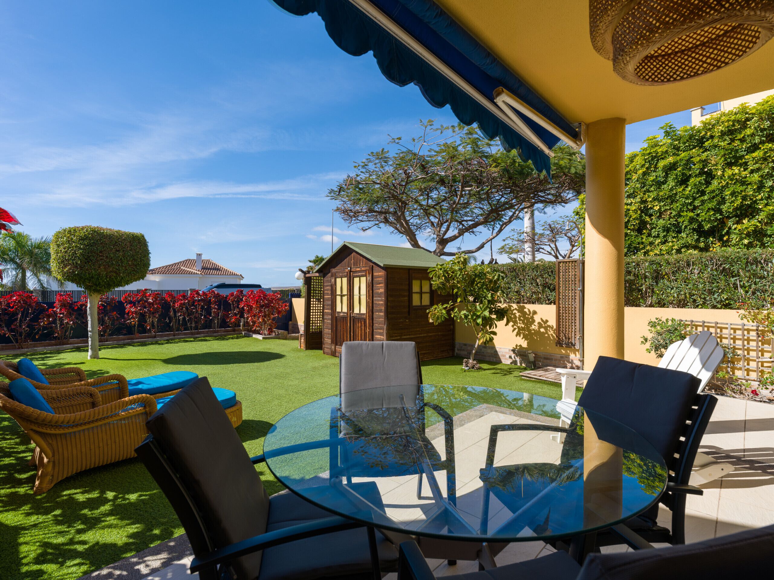 Een terras met een glazen tafel en stoelen, omringd door een verzorgd gazon en een klein houten gebouwtje.