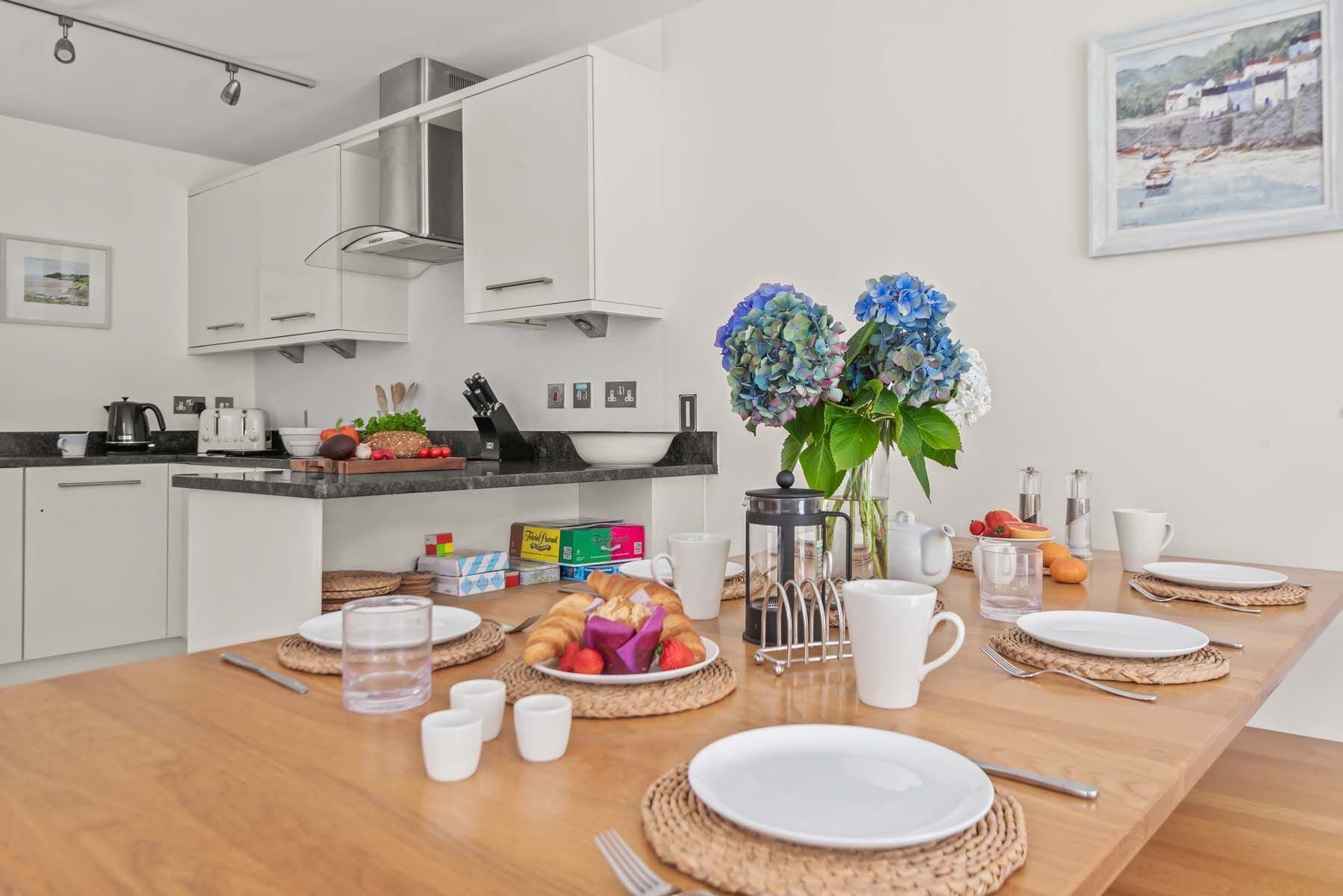 Une cuisine moderne avec des armoires blanches, une hotte en acier inoxydable et un coin repas aménagé pour le petit-déjeuner, avec des assiettes, des tasses et un vase de fleurs.