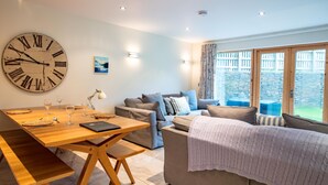 A modern living room with a large wall clock, a sofa, a dining table set with glasses, and a view of the outside. - House 1 - sleeps 6 guests  in 3 bedrooms (Looe)