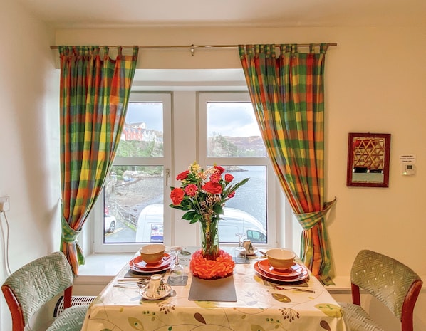 Mesa de comedor preparada con platos, tazones y un jarrón de flores, ubicada junto a una ventana con cortinas.