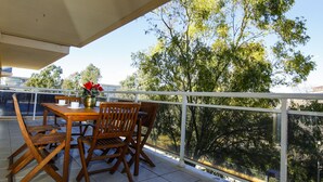 Un balcón con mesa y sillas de madera, un florero con flores y vistas a árboles y edificios.