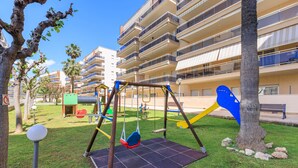 Un parque infantil con columpios y un tobogán, rodeado de césped y árboles, con un edificio al fondo.