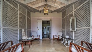A spacious room with a high ceiling, a chandelier, and a view into another room. There are wooden chairs and a table with a decorative item on it. - We Stay Well Sanctuary Barbados - Wellness in Paradise (Bridgetown)