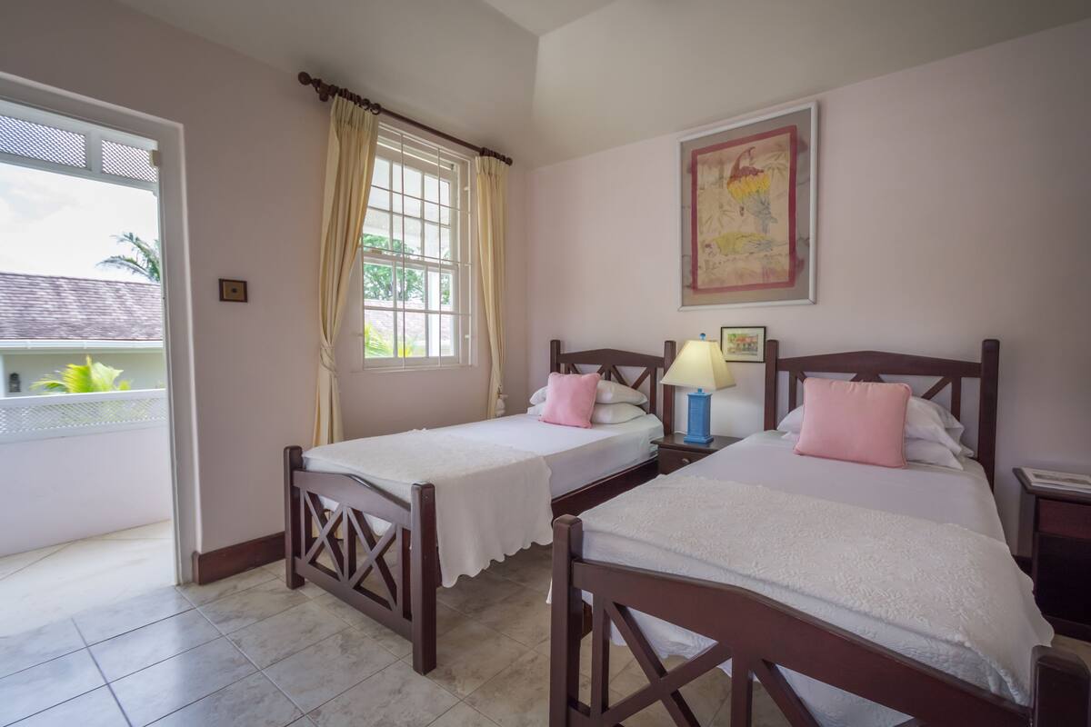 Una habitación con dos camas de madera, una ventana con cortinas, un cuadro enmarcado en la pared y una mesita de noche con una lámpara.