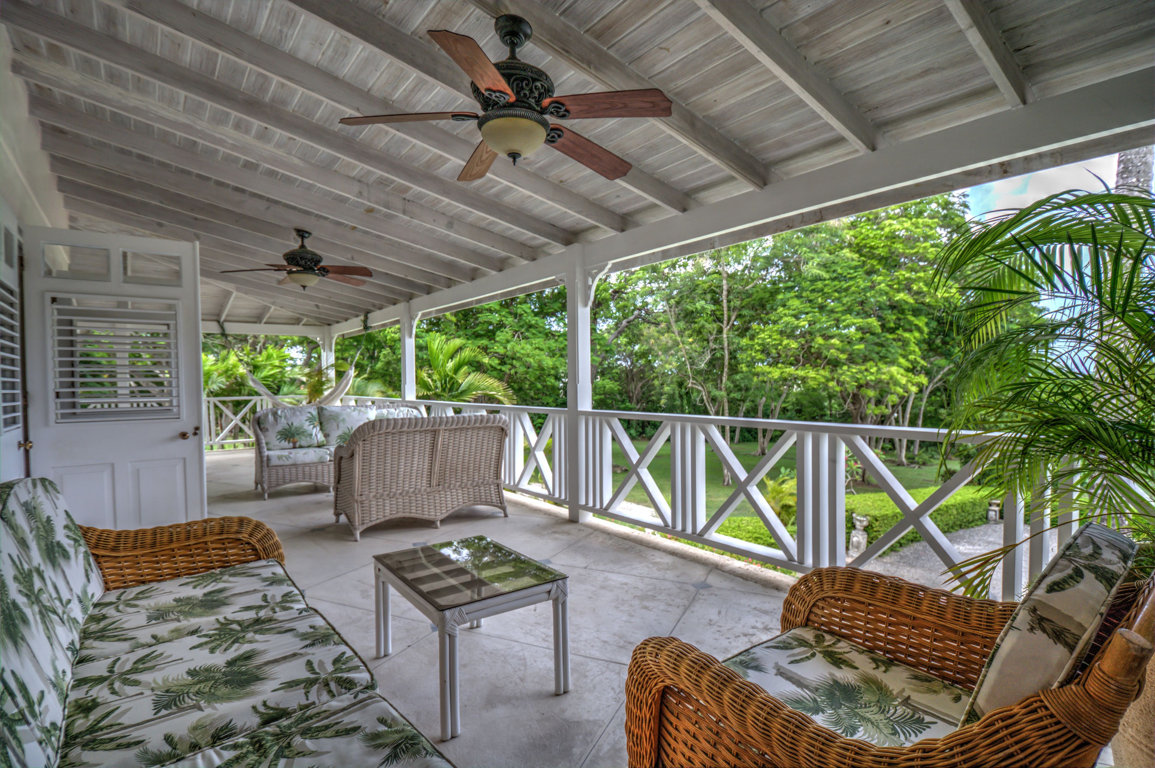 A covered porch with wicker furniture, ceiling fans, and a view of lush greenery.