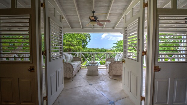 An open porch with wicker furniture and a ceiling fan, leading to a lush green garden. - We Stay Well Sanctuary Barbados - Wellness in Paradise (Bridgetown)
