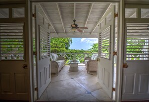 An open porch with wicker furniture and a ceiling fan, leading to a lush green garden. - We Stay Well Sanctuary Barbados - Wellness in Paradise (Bridgetown)