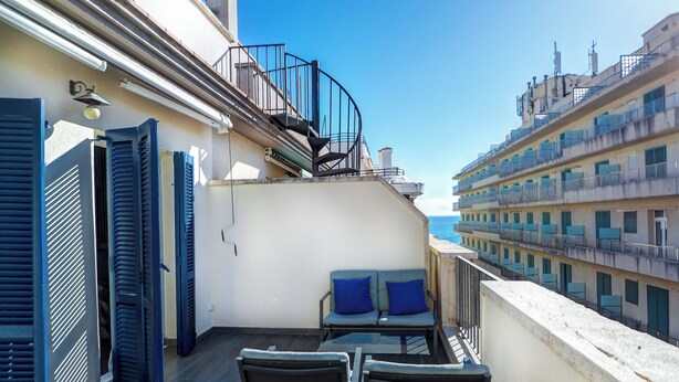 Un balcon avec deux chaises et une table, d’où l’on peut admirer un paysage urbain avec ses bâtiments et un ciel dégagé.