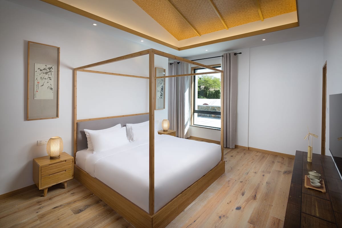 Une chambre à coucher moderne avec un lit à baldaquin, des tables de chevet en bois, une lampe de chevet et une vue sur l’extérieur.