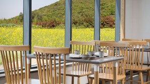 Een eetruimte met houten stoelen en tafels, grote ramen met uitzicht op een groene heuvel en gele bloemen.