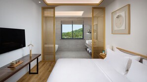 Een moderne hotelkamer met een groot bed, een televisie, een houten bankje, een raam met uitzicht op groen en een kunstwerk in een lijst aan de muur.