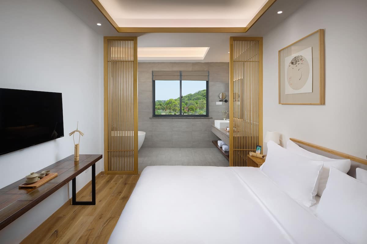 Une chambre d’hôtel moderne avec un grand lit, une télévision, un banc en bois, une fenêtre donnant sur un paysage verdoyant et une œuvre d’art encadrée au mur.