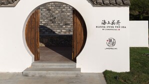 Een modern gebouw met een gevel van steen, een houten deur en een bord met de tekst 'BLOOM OVER THE SEA BY JINSPECIAL'.