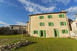 A stone building with multiple windows and a door, surrounded by a grassy area and a stone wall.