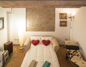 A bedroom with a brick wall, a bed with white bedding, two heart-shaped pillows, a side table, a lamp, and a chair with a cowhide pattern.