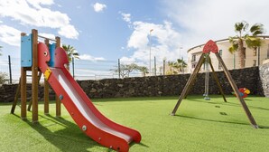 A playground with a red slide and swings on artificial grass, surrounded by a stone wall and palm trees. - Adeje with pool, sea view and WIFI gratis (Adeje)