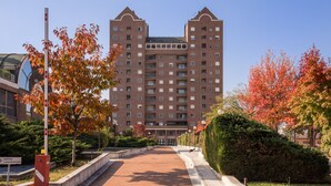 Un edificio de varios pisos con fachada simétrica, entrada central y un área ajardinada con árboles y un camino pavimentado.