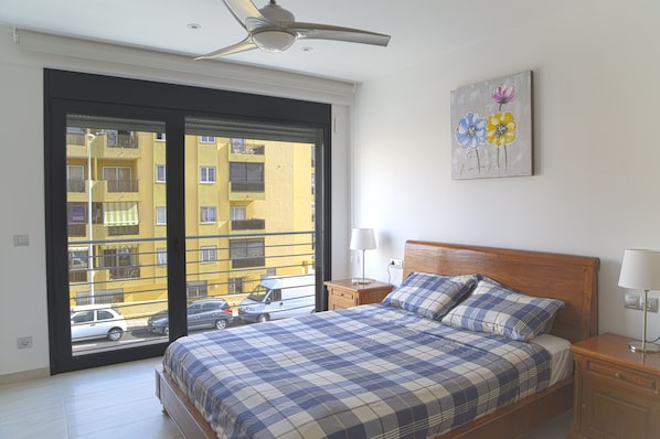 A bedroom with a large bed, a ceiling fan, a nightstand, a lamp, and a window with a view of a yellow building and parked cars.