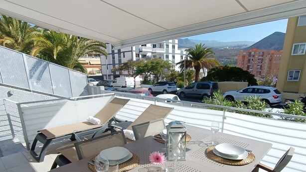 An outdoor dining area with a table set for four, surrounded by chairs, on a terrace with a view of buildings and parked cars. - Idaira III new spacious apartment with private garage and beach walking distance (Los Cristianos)