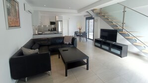 A modern living room with a black leather sofa, a coffee table, and a TV stand. There is a kitchen in the background with white cabinets and a stainless steel refrigerator. A staircase with glass railings leads to an upper level. - Idaira III new spacious apartment with private garage and beach walking distance (Los Cristianos)