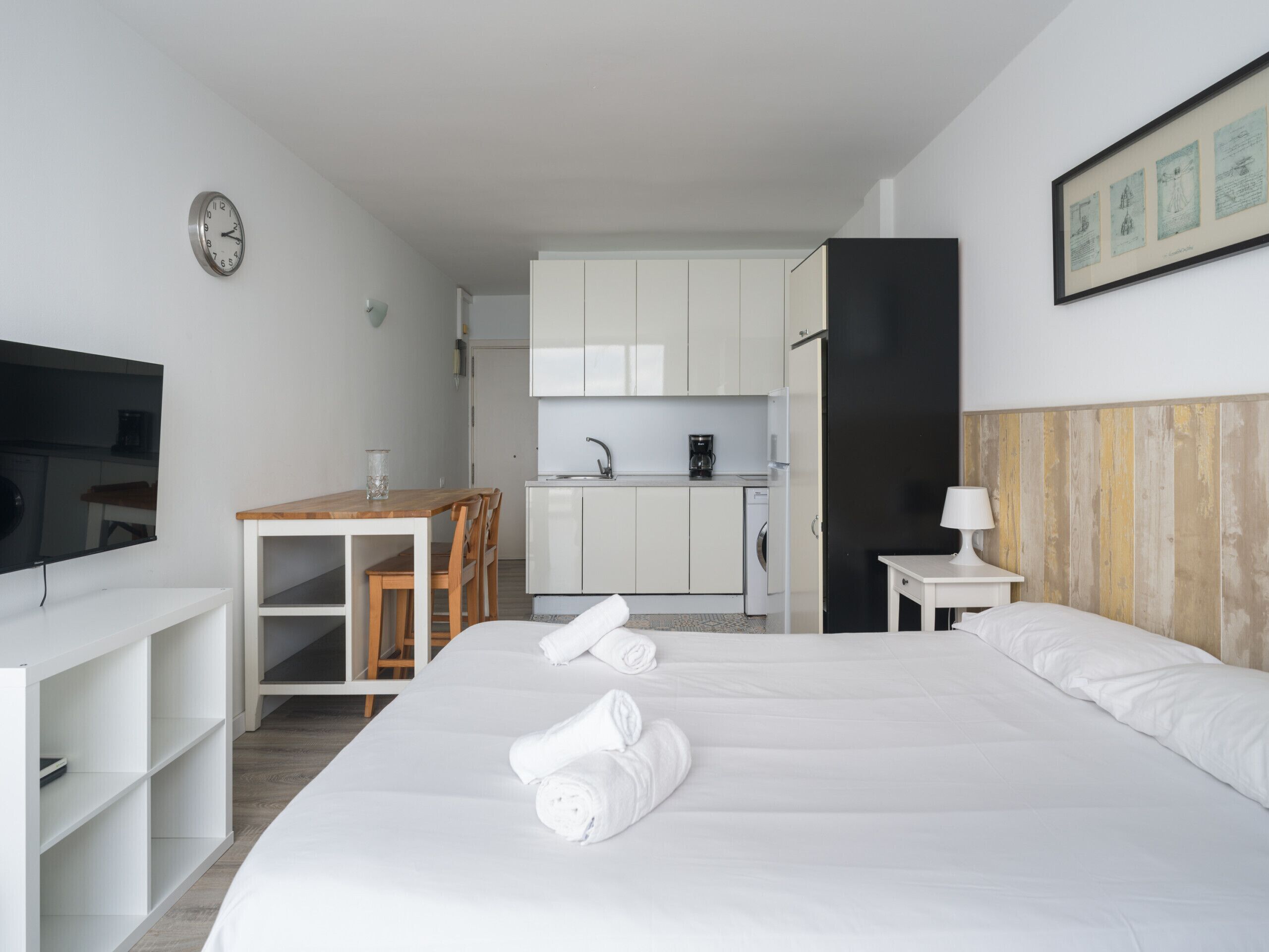 A modern hotel room with a large bed, a desk, a kitchenette, and a TV.