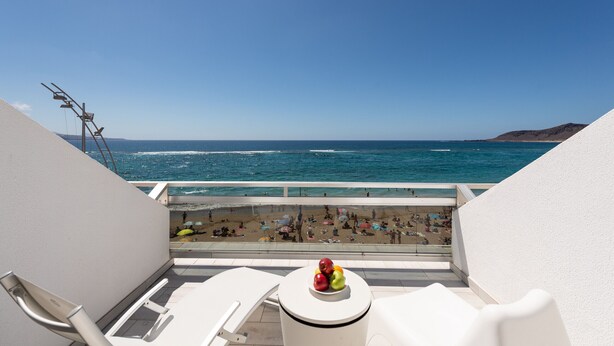 Varanda com vista para a praia e o oceano. Há cadeiras brancas e uma pequena mesa com frutas.