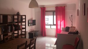 A living room with a wooden table, chairs, a bookshelf, a TV, and a window with red curtains. - FAMILY APARTMENT LISBON - TAGUSPARK (São Marcos)