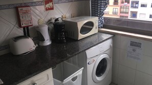 A kitchen with a microwave, toaster, and coffee maker on the counter, a washing machine, and a window with a view of buildings. - FAMILY APARTMENT LISBON - TAGUSPARK (São Marcos)