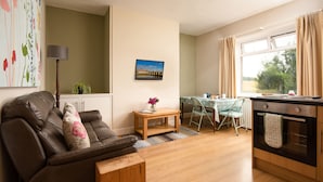 A living room with a brown leather sofa, a wooden coffee table, a dining area with a table and chairs, and a kitchen with an oven.