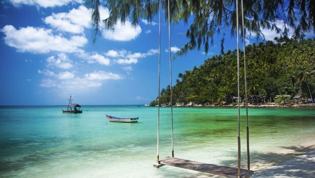 Ein Strand mit klarem türkisblauem Wasser, einer Schaukel und einem Boot in der Ferne.