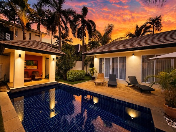 A swimming pool with a tiled deck, surrounded by palm trees and a house with a tiled roof.