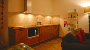 Een compacte keuken met houten kastjes, een ingebouwde oven en een aanrecht. Er is een zithoek met een donkergrijze bank en een lamp.