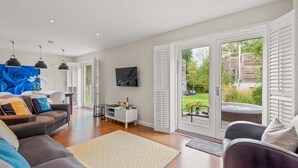 Ein geräumiges Wohnzimmer mit einer Couch, einem Couchtisch und einem Fernseher. Es gibt Glastüren, die zu einer Terrasse mit Whirlpool und Garten führen.