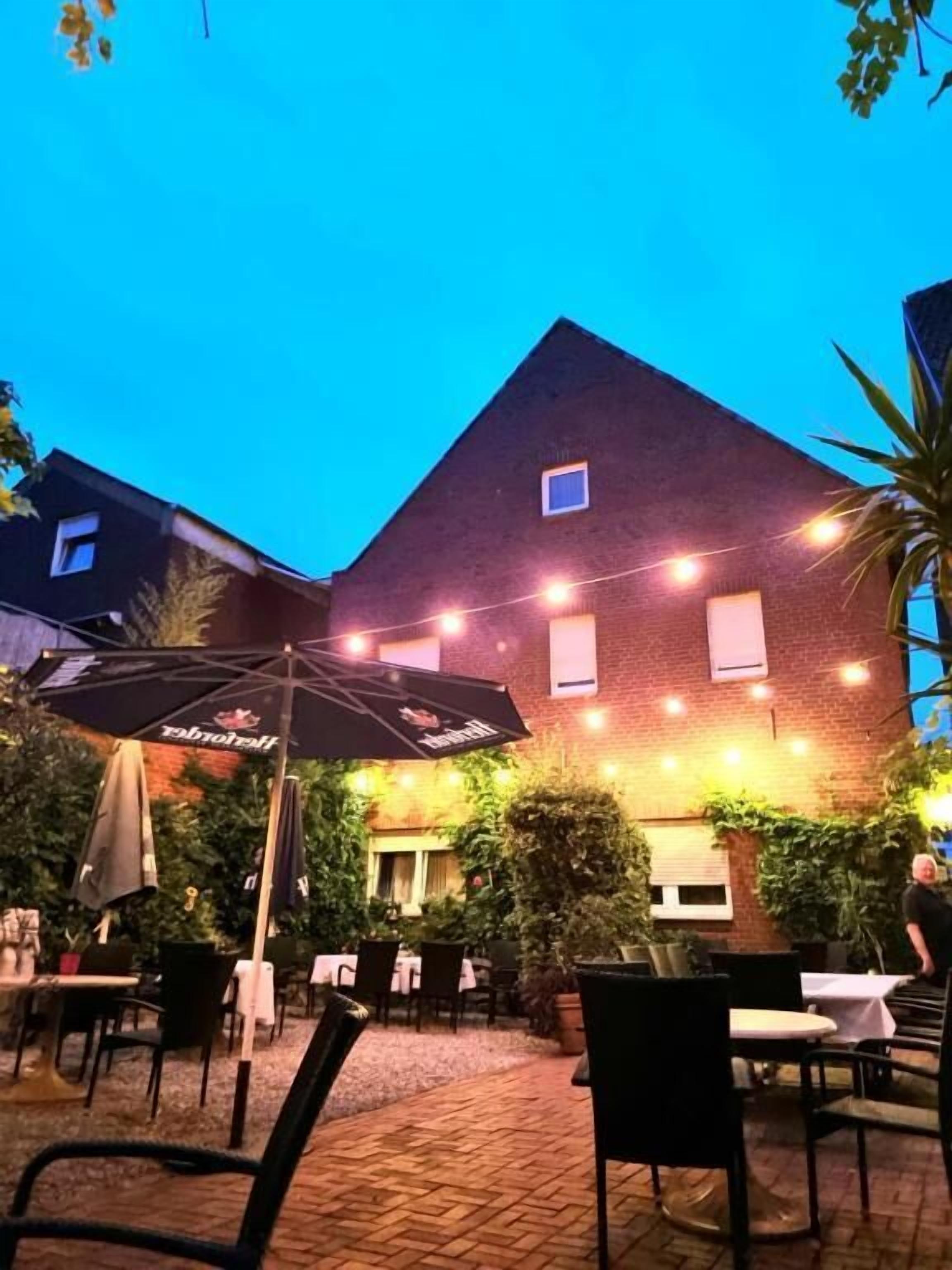 Ein Außenbereich mit Tischen und Stühlen, ein Backsteinhaus im Hintergrund und Lichterketten.