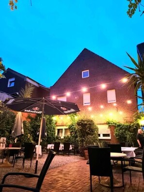 An outdoor seating area with tables and chairs, a brick building in the background, and string lights. - Hotel Altes Gasthaus Düsterbeck (Emsdetten)