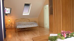 A bedroom with a bed, a wooden wardrobe, a small table with flowers, a ceiling fan, a window with blinds, and a TV on a stand.