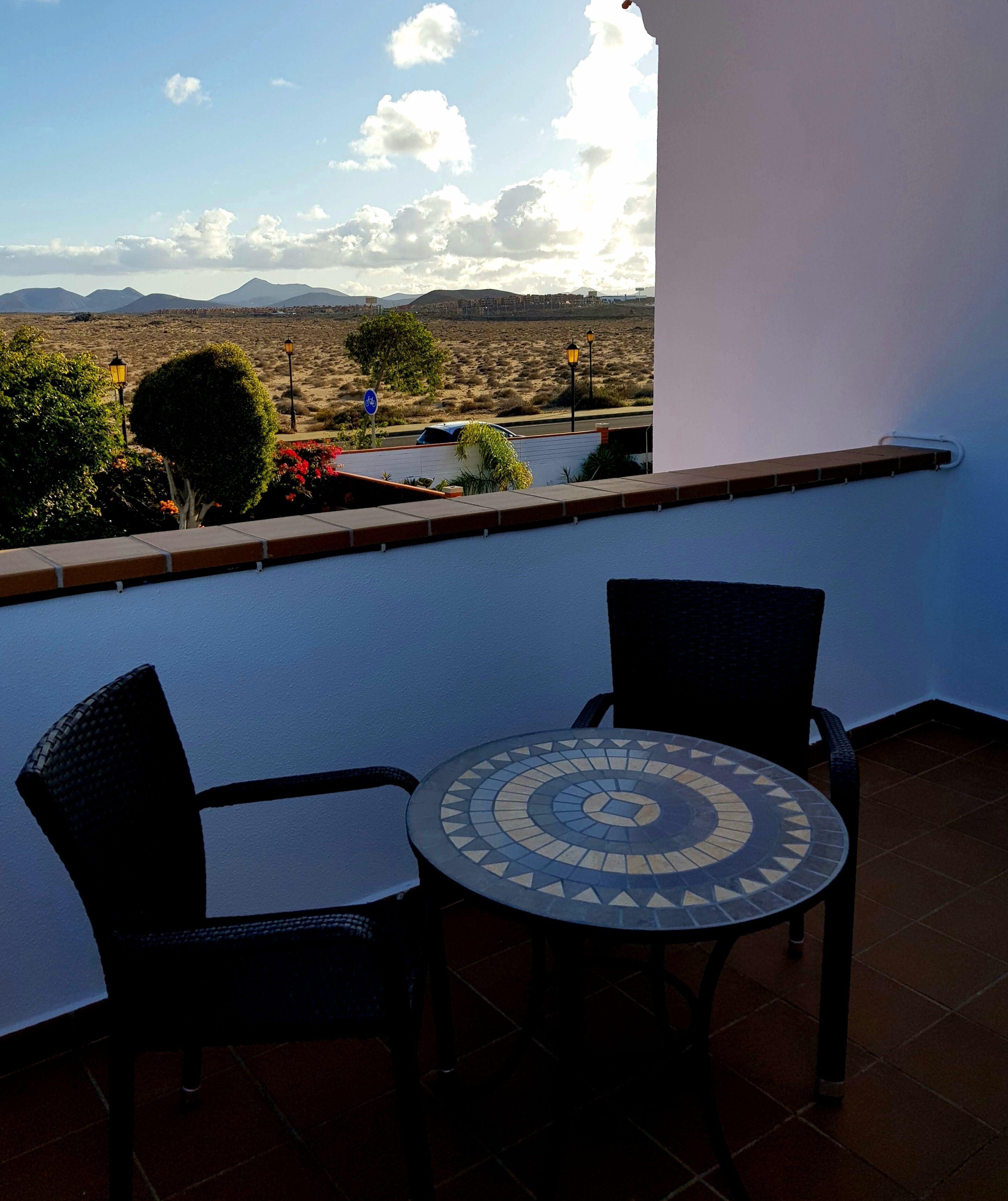 A balcony with two chairs and a table, overlooking a desert landscape with mountains in the distance.