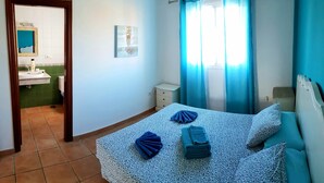 A bedroom with a bed, a nightstand, a window with curtains, a bathroom visible through an open door, and a wall-mounted picture. - Wonderful villa with a large private heated pool and stunning views to the dunes (Corralejo)