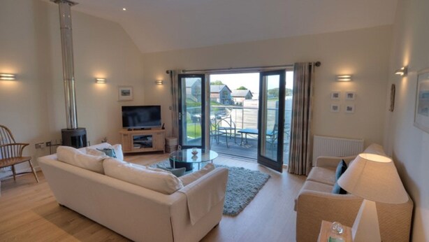 A modern living room with a sofa, TV, and a fireplace. There is a glass door leading to an outdoor area with a table and chairs. - Beautiful holiday home, just a walk away from Talland beach... (Looe)