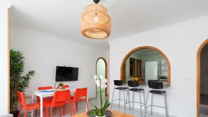 A modern living space with a dining area, a TV mounted on the wall, and a kitchen visible through an archway.