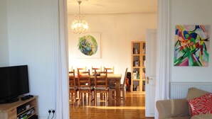 Een woonkamer met een televisie, houten meubels, een witte bank, een rode kussen, een boekenkast en een schilderij aan de muur.