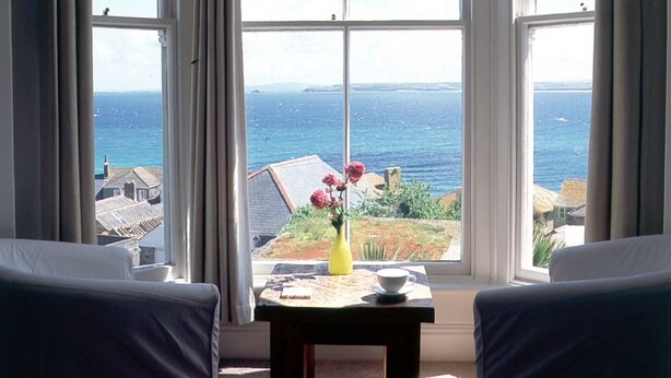 Een kamer met een groot erkerraam met uitzicht op zee, een kleine tafel met een bloemetje en twee stoelen.