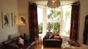 Een gezellige woonkamer met een bruine leren bank, een zacht, wit vloerkleed en een relax fauteuil. Twee ramen met rode gordijnen, een radiator en wanddecoratie.