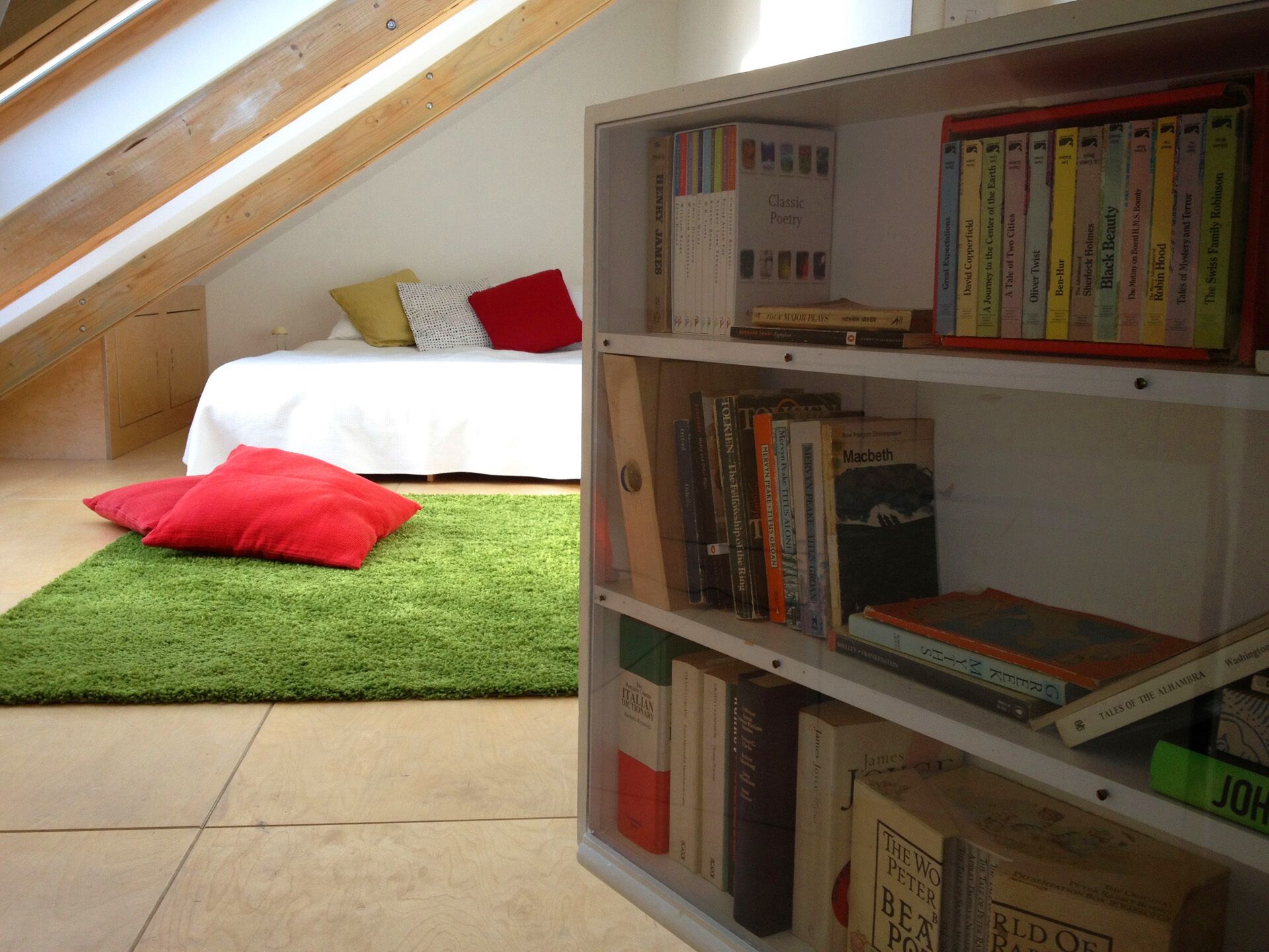 Una habitación con techo inclinado, una cama con almohadas, un tapete verde y una estantería con varios libros.
