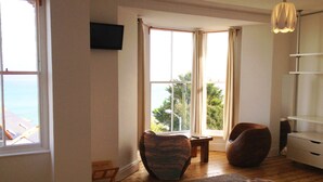 Een slaapkamer met een groot raam, een bed, een stoel, een klein tafeltje en een aan de muur bevestigde televisie.