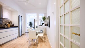 A modern kitchen with a stainless steel refrigerator, a dining area with a glass table and white chairs, and a large window with a decorative glass door.